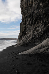 Distinctive basalt columns form a natural cliff face on a stark black sand beach with footprints...