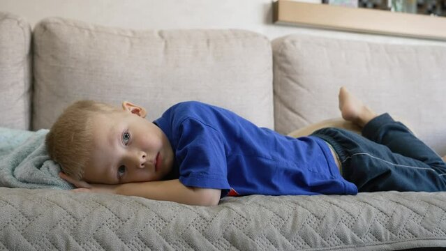 Cute bored child watching TV while lying on the sofa in the living room. Lazy lifestyle. A blond boy spends time lying on the sofa during the weekend. Domestic life of real people.