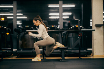 A fit sportswoman is doing split squats on a bench in a gym.