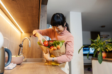 A japanese middle-aged woman is holding shopping bag with vegetables in kitchen.