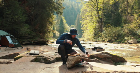 Close-up of a lone tourist sitting on a stone in a tourist camp near a fire and chopping wood