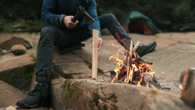 Close-up of a lone tourist sitting on a stone in a tourist camp near a fire and chopping wood