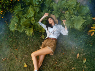 A woman in a hat and shorts laying on the grass with smoke coming out of her mouth