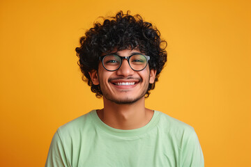 Happy millennial hindu guy in eyeglasses and bright green t-shirt with bushy curly hair posing alone on yellow studio background, cheerfully smiling at camera, closeup portrait