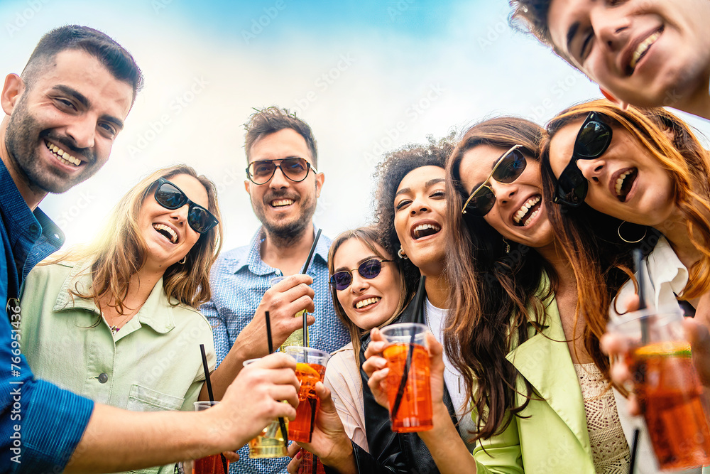 Wall mural Laughing group of friends toasting with cocktails - celebrating friendship, summer joy, carefree outdoor fun - young adults with drinks in plastic glasses - copy space in the sky