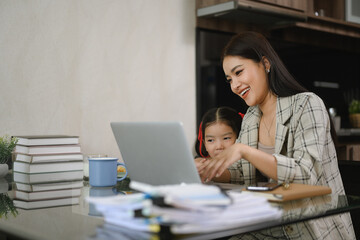 Smiling mother working remotely with laptop sitting with her cute little daughter in cozy kitchen