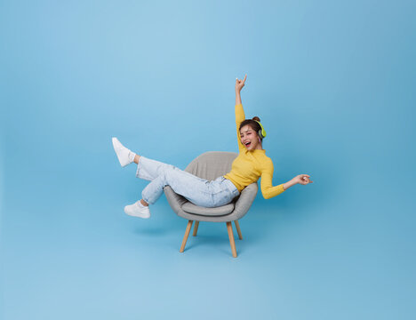 Happy young asian girl listening music with headphone dancing on chair isolated on blue background. People lifestyle concep