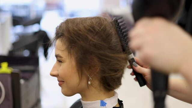 happy woman with short brunette hair sitting in hairdressing cape in beauty salon, getting haircut by professional hairdresser.girl doing keratin treatment.curls at the end.wet hair,towel on head.4k