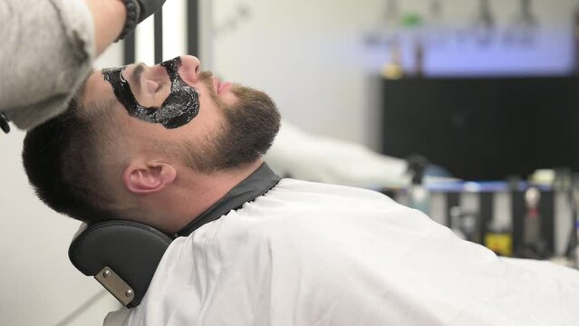 Barber applies a black moisturizing mask to a man's face while shaving. A dermatologist applies a black mask to the face of a bearded patient to clear pores and wrinkles.