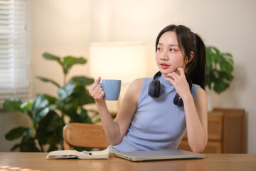 Young attractive happy Asian female student holding a cup of coffee sitting in the living room smiling and looking at the left while working on a laptop at the home office.