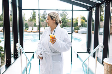 A young woman in a white bathrobe smiles and stands in the spa salon. A charming girl is drinking orange juice by the pool.