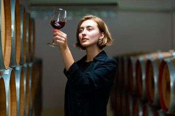 Professional Caucasian woman sommelier tasting and sniffing red wine in wine glass at wine cellar with wooden barrel. Winery brewery liquor wine factory manufacturing industry and winemaker concept.  - obrazy, fototapety, plakaty