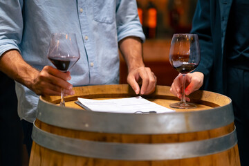 Professional man and woman sommelier tasting and smelling red wine in wine glass at wine cellar with wooden barrel at wine factory. Alcohol liquor shop, brewery, winery industry and winemaker concept.
