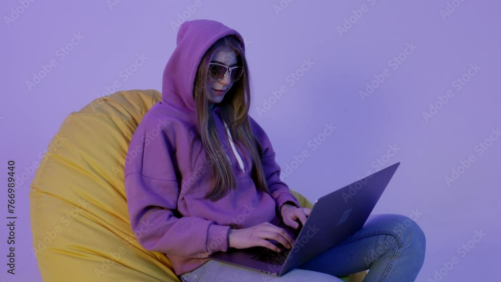 Wall mural young woman sitting on pouf and using notebook isolated on violet color background. girl in hoodie w