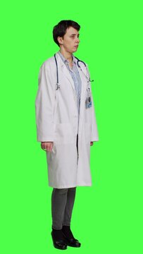 Side view Physician saying no and showing negative reaction symbol in studio, wearing a white coat and standing against greenscreen backdrop. Woman medic feeling displeased, rejection sign. Camera A.