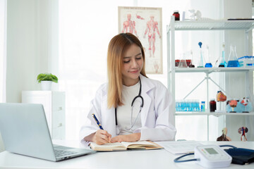 Obraz na płótnie Canvas Young Asian woman or female medical staff sitting smiling, arms looking happily at camera, working on table in clinic office in telehealth, telemedicine, consulting online service.