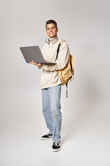 young student in headphones standing with backpack and networking to laptop against grey background