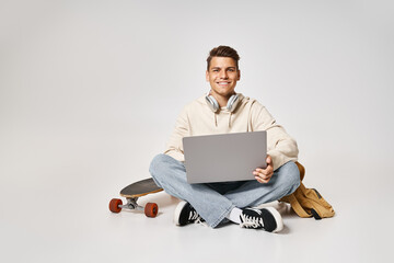 smiling young student in headphones networking to laptop and sitting with backpack and skateboard