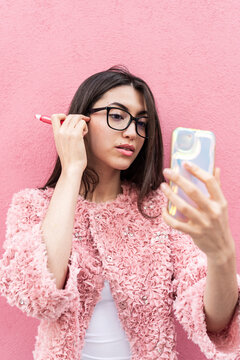 Moroccan woman using smartphone to adjust eyewear against pink wall, makeup fine-tuning