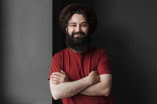 Portrait of a cheerful bearded man with a friendly smile, wearing a red t-shirt and a stylish hat, confidently posing with arms crossed over a gray background.