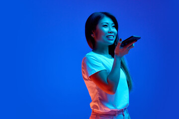 Beautiful Asian woman using phone and looking away against blue gradient background in neon light....