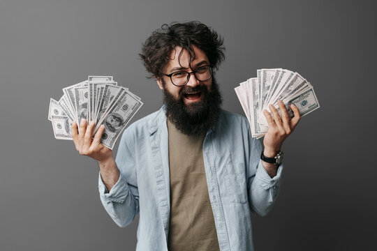 A casually dressed man stands against a neutral background, holding a wad of cash with both hands. The image conveys concepts of wealth, finance, savings, and success without showing the man's face.