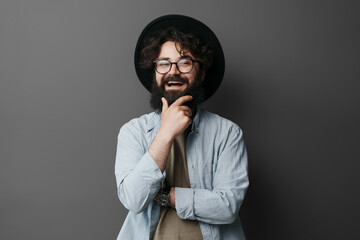 Portrait of a cheerful bearded man wearing glasses and a stylish hat, confidently posing with a hand on his chin against a grey studio background, expressing individuality and fashion sense. - obrazy, fototapety, plakaty