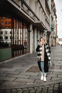 Stylish Woman with Coffee Cup Strolling Down Street. Chic woman wearing scarf enjoys a leisurely walk down the street while holding a coffee cup. Urban lifestyle and coffee on-the-go concept