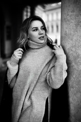 Naklejka premium Stylish woman in a gray sweater, black coat, and scarf poses confidently beside a pillar, exuding elegance and urban chic vibes. A woman in a gray sweater stands in front of a pillar.