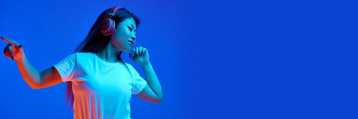 Banner. Asian woman listening music in headphones against blue gradient background in neon light....