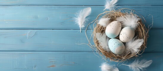 Easter eggs and feathers in a nest are displayed on a blue wooden surface in a simple and minimalistic setup. The image is captured from a top-down perspective with a card featuring space for text. - obrazy, fototapety, plakaty