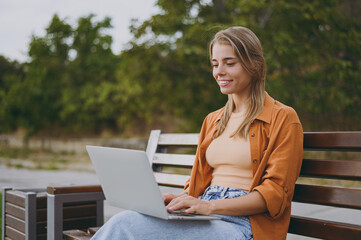 Side view young IT woman wearing orange shirt casual clothes hold use work on laptop pc computer sit on bench walking rest relax in spring green city park outdoors on nature. Urban lifestyle concept.