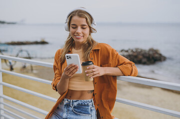 Young smiling woman wears orange shirt casual clothes headphones listen to music use mobile cell phone drink coffee walk on sea ocean sand shore beach outdoor seaside in summer day. Lifestyle concept. - 767029054