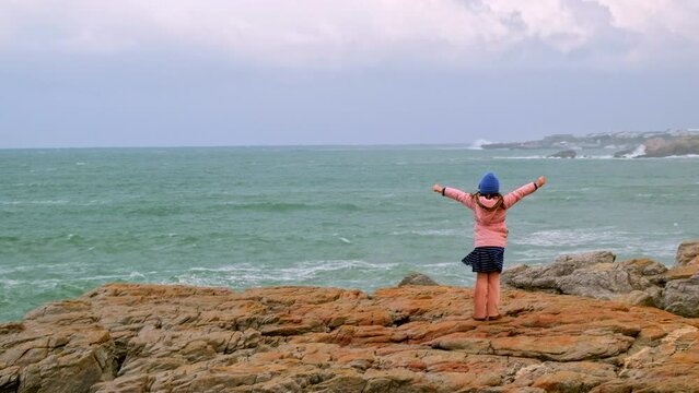 Brave young girl stands with outstretched arms looking at ocean as massive wave crashes against rocky shore in front of her and she retreats