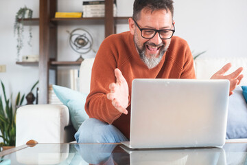 Adult Caucasian man wearing glasses sit on sofa at home talk on video call on laptop, freelance male working online have webcam virtual online conference conversation on computer with client remote - obrazy, fototapety, plakaty