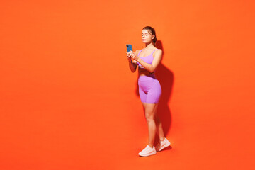 Fototapeta na wymiar Full body young fitness trainer instructor woman sportsman in top shorts purple clothes train in home gym hold use mobile cell phone isolated on plain orange background Workout sport fit abs concept