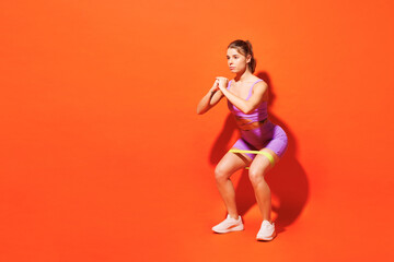 Fototapeta na wymiar Full body young fitness trainer woman sportsman wearing purple top clothes in home gym using elastic rubber band for legs do squats isolated on plain orange background. Workout sport fit abs concept.