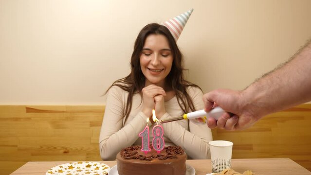 The girl is sitting in front of a table with a festive cake, in which a candle in the form of the number 18 is burning, which she blows out. Birthday celebration concept.
