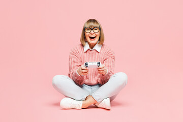 Full body elderly woman 50s years old wear sweater shirt casual clothes glasses sits hold in hand play pc game with joystick console isolated on plain pastel light pink background. Lifestyle concept.