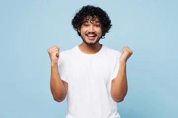 Gardinen Young fun happy Indian man he wear white t-shirt casual clothes doing winner gesture celebrate clenching fists isolated on plain pastel light blue cyan background studio portrait. Lifestyle concept. © ViDi Studio