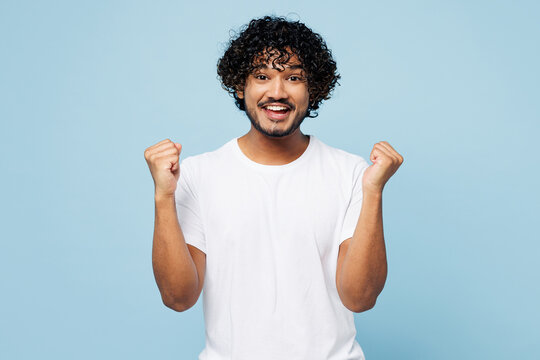 Young fun happy Indian man he wear white t-shirt casual clothes doing winner gesture celebrate clenching fists isolated on plain pastel light blue cyan background studio portrait. Lifestyle concept.