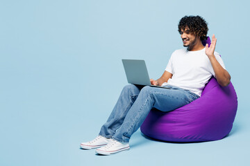 Full body young happy Indian man wear white t-shirt casual clothes sit in bag chair hold use work on laptop pc computer waving hand isolated on plain pastel light blue background. Lifestyle concept