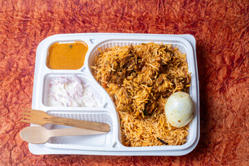Indian chicken biryani in white plastic box filled with raita and gravy and wooden spoon on wooden table.