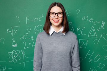 Young smiling happy cheerful smart teacher woman she wear grey casual shirt glasses looking camera isolated on green wall chalk blackboard background studio. Education in high school college concept. - obrazy, fototapety, plakaty