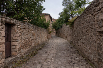 Fototapeta na wymiar A narrow, stone-paved street flanked by old, textured stone walls, leading to aged buildings, under an overcast sky