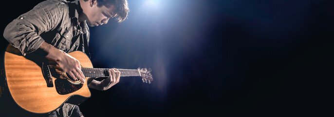 A male guitarist plays an acoustic guitar on a dark stage background.