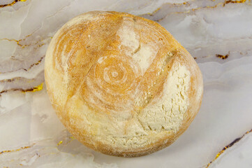 A bread with a brown crust sits on a marble countertop. The bread appears to be freshly baked and has a slightly rough texture. Concept of warmth and comfort