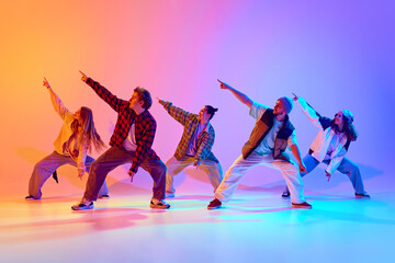 Fototapeta premium Group of five dancers in casual clothes performing with synchronized poses against gradient studio background in neon light. Concept of modern dance style, hobby, active lifestyle, youth culture