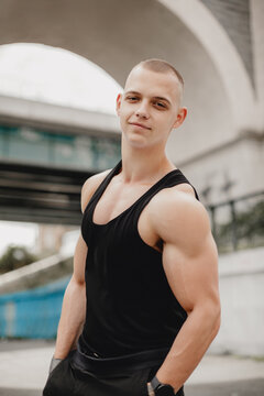 A man with a muscular build stands in front of a bridge, wearing a black tank top and shorts. He is posing for a photo, with his hands in his pockets. Concept of confidence and strength