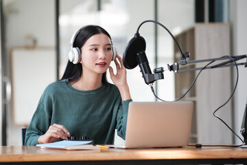 A woman wearing headphones is sitting at a desk with a laptop and a microphone. She is recording a...